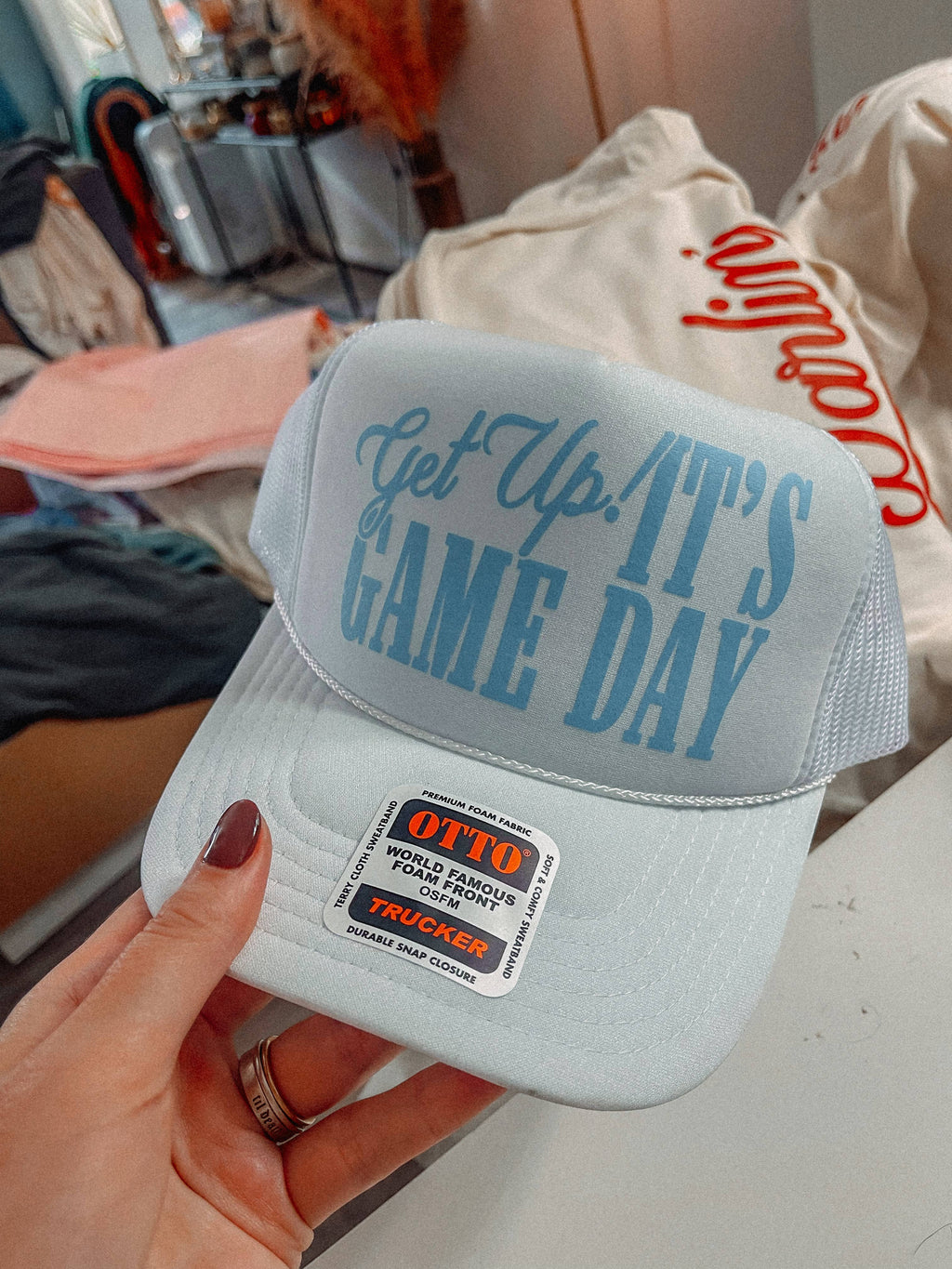 White cap with 'Get Up! It's Game Day' text held by a hand in an indoor setting.