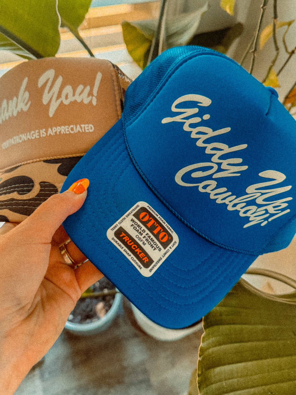 Two hats, one beige with 'Thank You!' text and the other blue with 'Giddy Up!' text, held by a hand against a plant background.