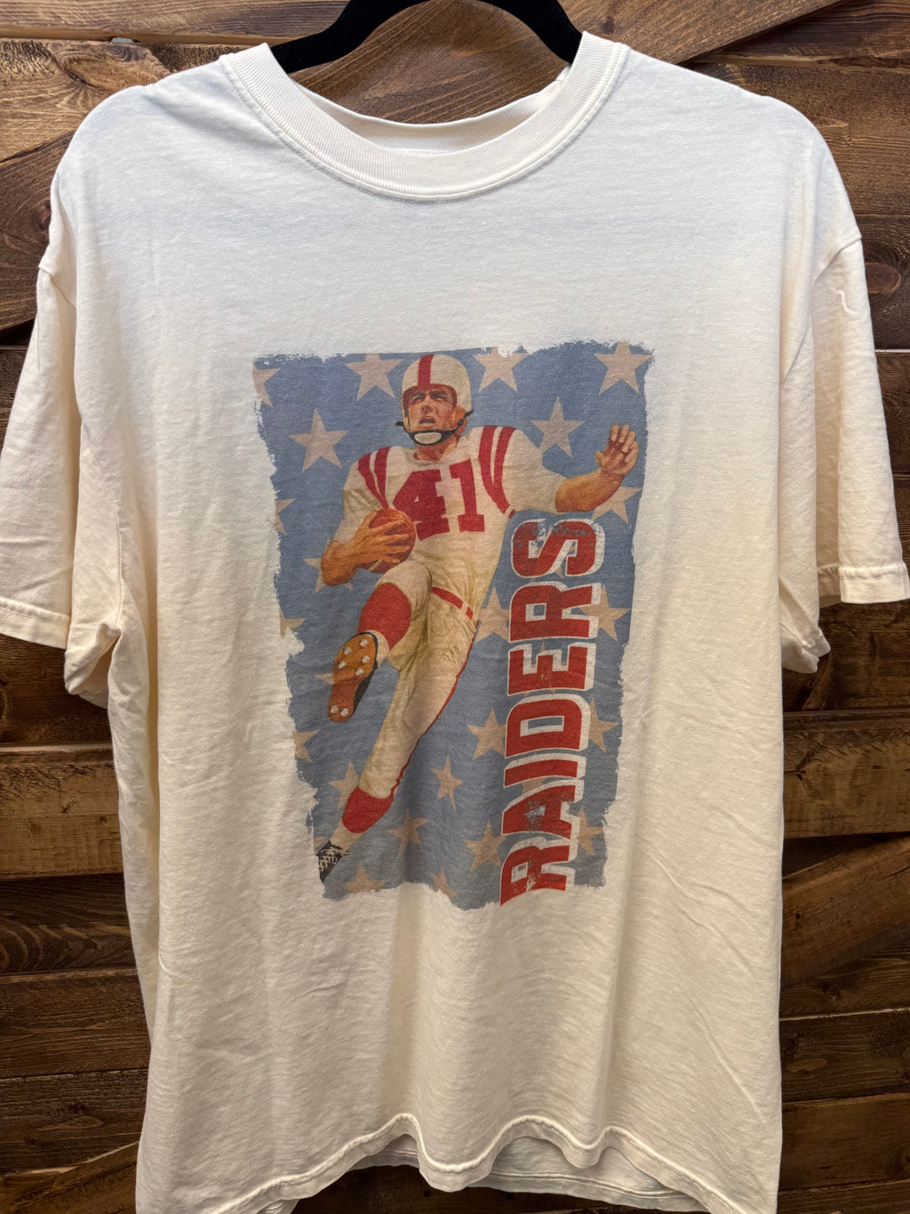 A football-themed clothing display featuring a cream graphic t-shirt with a football player and 'RAIDERS' text, styled with blue and white striped shorts, a red cap, and a blue cap on a wooden display stand.