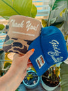 Two hats, one beige with 'Thank You!' text and the other blue with 'Giddy Up!' text, held by a hand against a plant background.