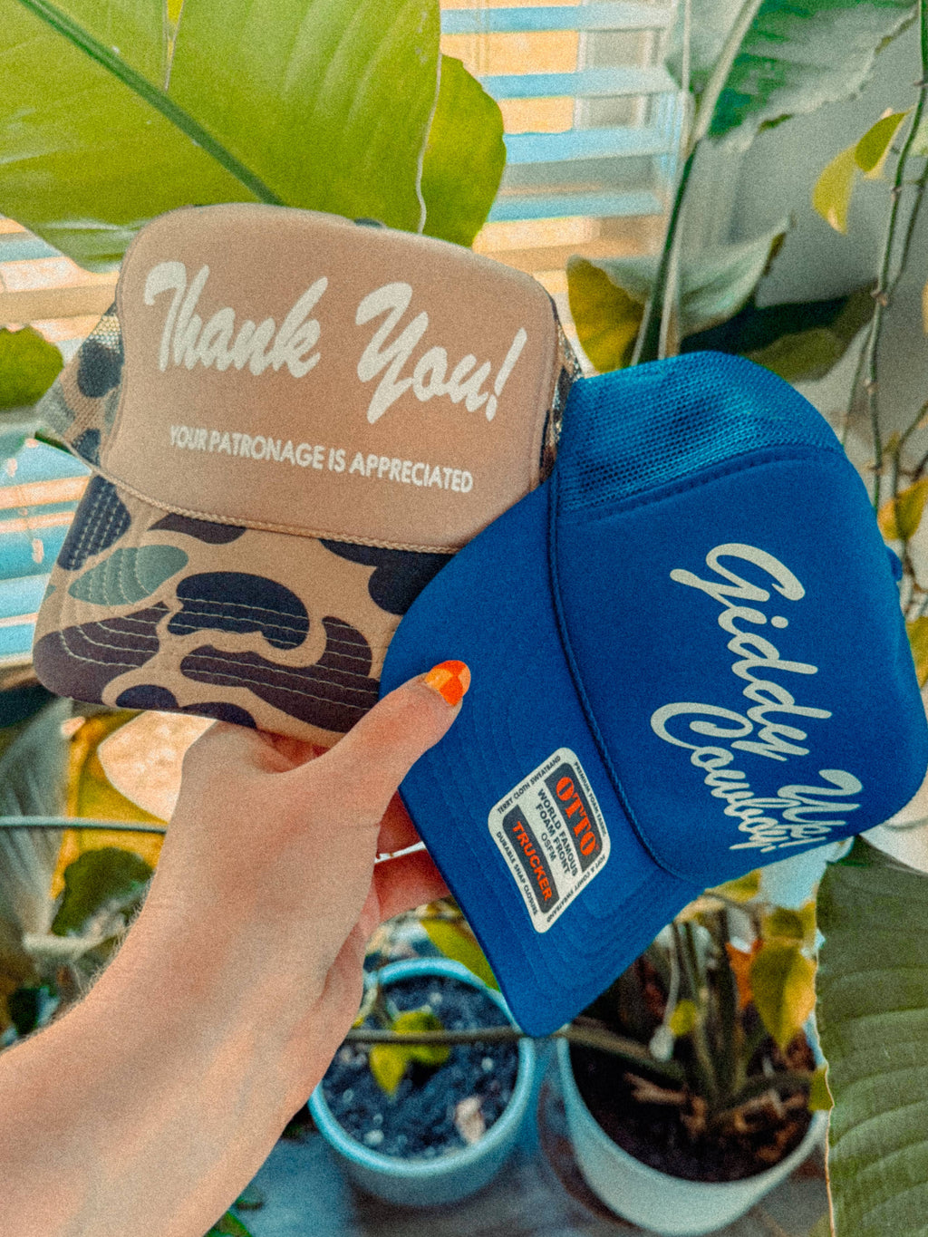 Two hats, one beige with 'Thank You!' text and the other blue with 'Giddy Up!' text, held by a hand against a plant background.